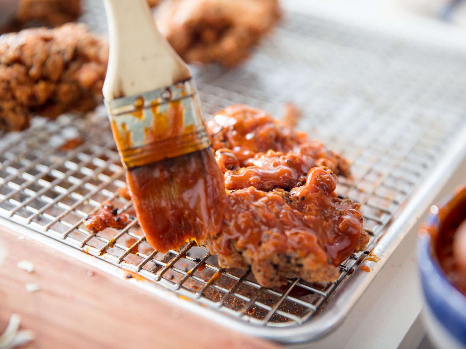 A brush swipes a Korean-inspired hot sauce over a piece of fried chicken on a wire rack.