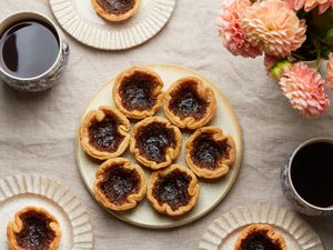 Overhead of platter with Canadian butter tarts. 3 smaller plates have one tart each. Coffee and flowers on the tabletop, with linen tablecloth.