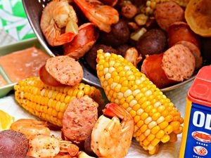 Closeup of a colander full of low-country shrimp boil on its side, cornucopia-like, with shrimp, halved corn cobs, and chunks of sausage spilling forth. Old Bay seasoning and dipping sauce are visible on either side.