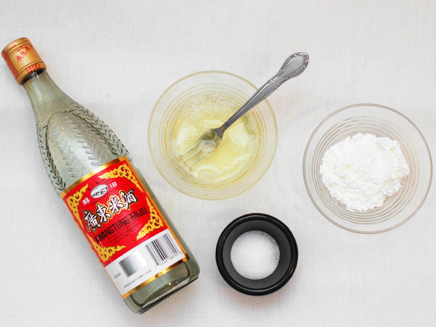 The ingredients for the velveting marinade on a white surface: rice wine, a bowl of egg white, a bowl of cornstarch and a smaller bowl of salt. 