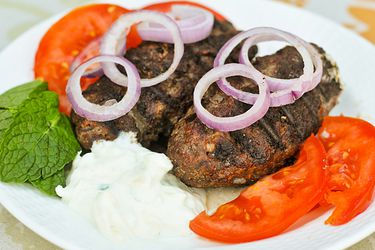 Greek bifteki on a plate with tomato slices, red onion slices, mint leaves, and tzatziki.