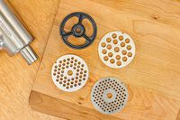 Different sizes of grinding plates of KitchenAid KSMMGA Metal Food Grinder Attachment displayed on wooden board