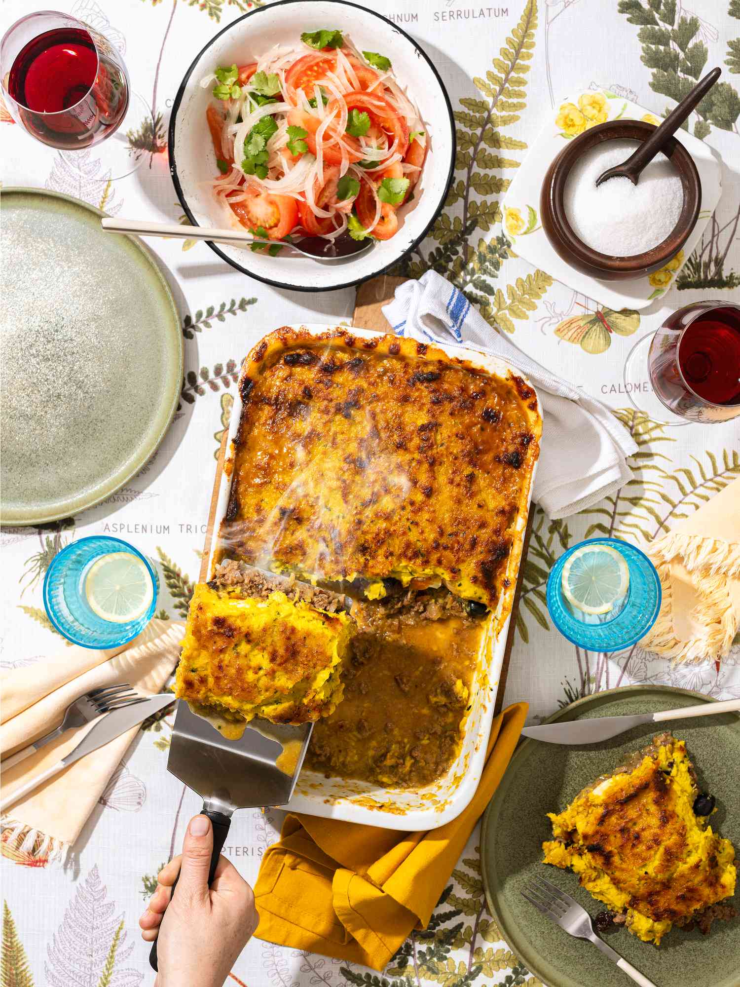 Overhead view of serving of pastel de choclo being picked up on a spatula, with another serving already on a plate. The table has another plate, wine, water, salt and salad