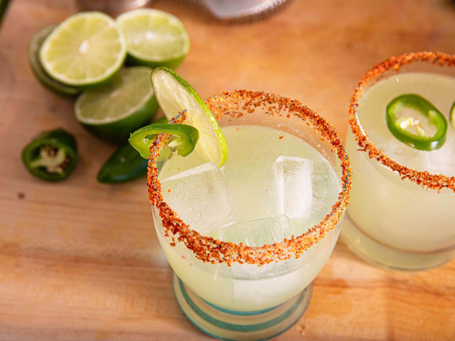 Two margarita glasses garnished with lime and chili flakes, with lime slices and a jalapeño in the background
