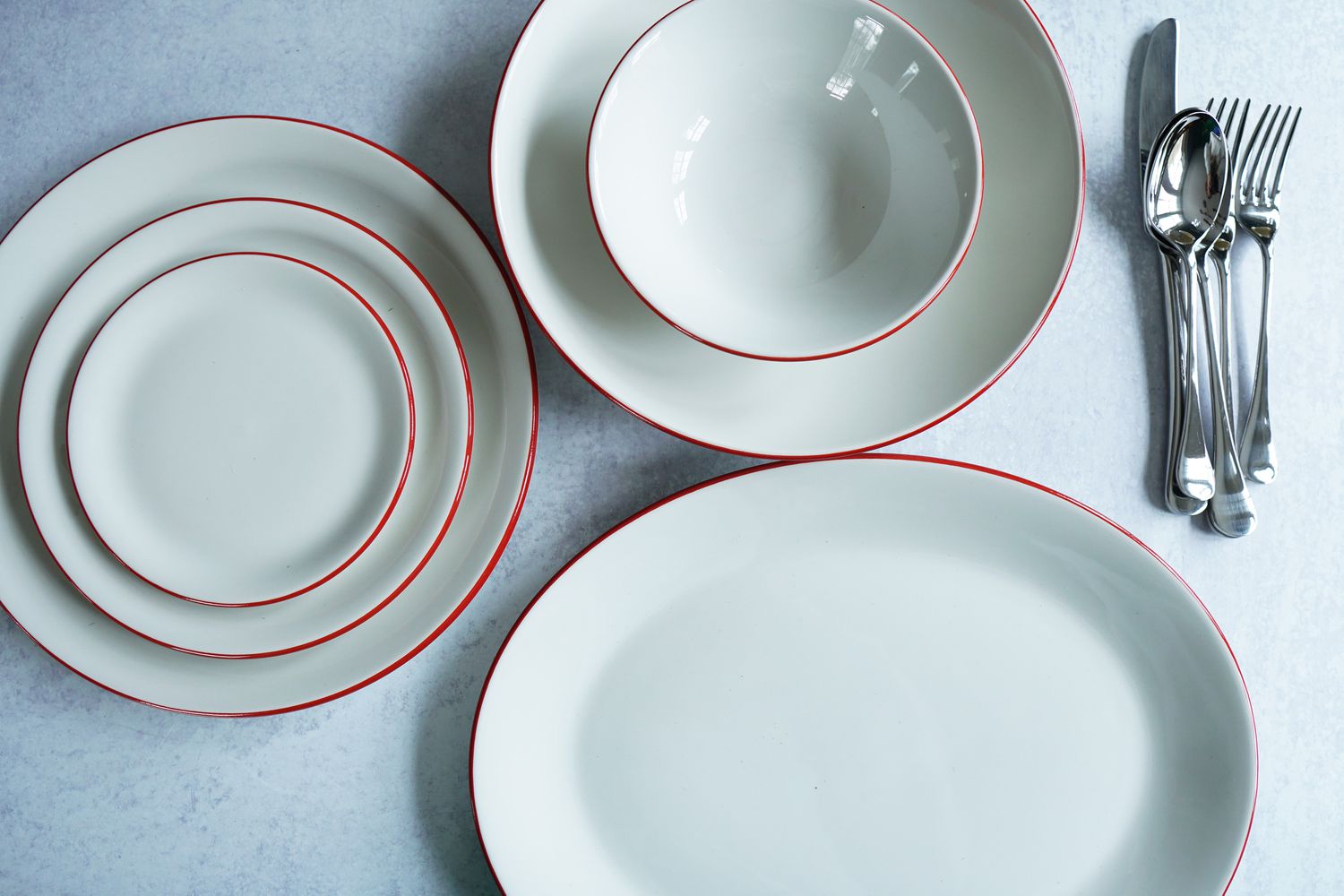 A white place setting with red trim, serving platter, and silverware on a grey surface