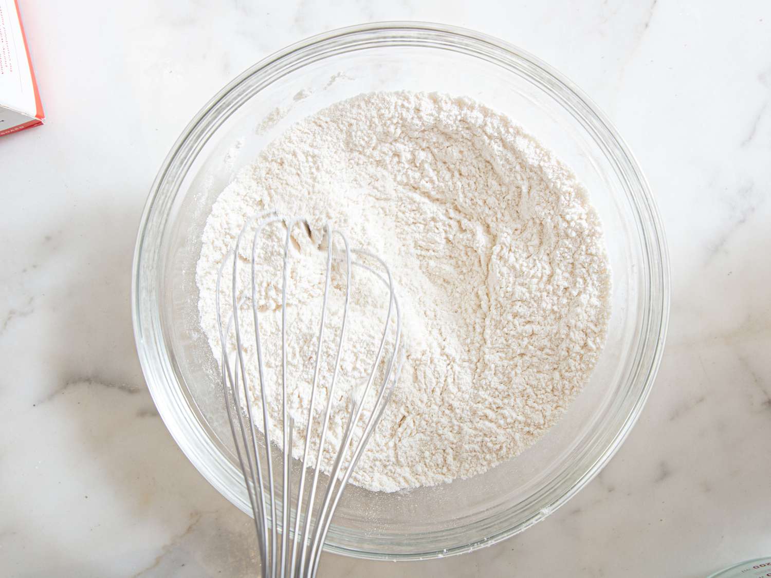 A whisk with a bowl of flour on a marble surface