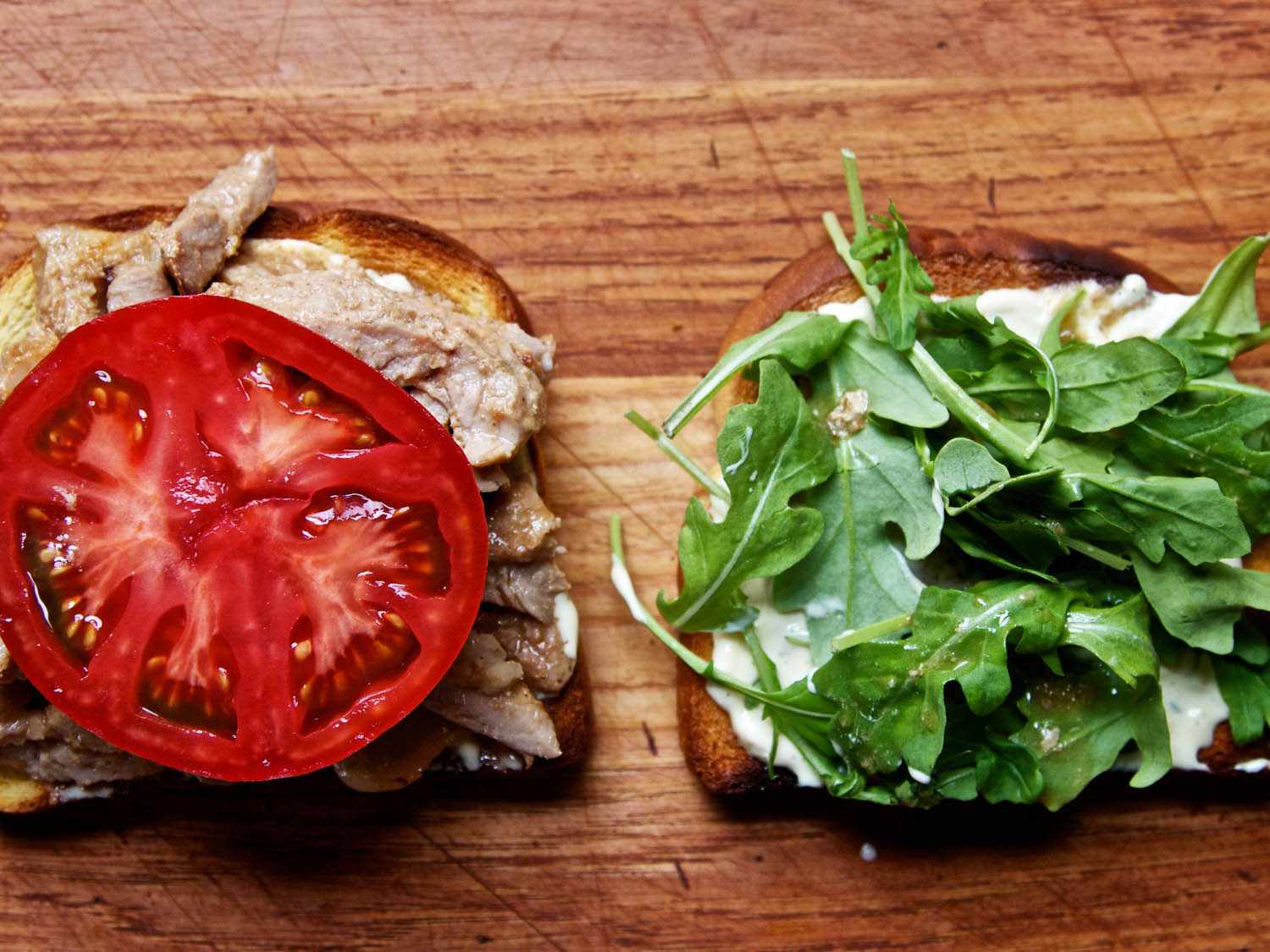 Overhead view of the sandwich before final assembly. A large slice of tomato and a small pile of arugula crown each slice of bread.