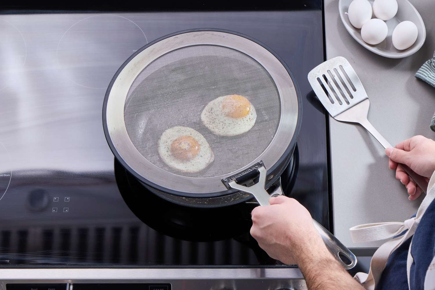 a person holding the le creuset splatter screen over a skillet with fried eggs.