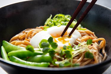 A bowl of mazemen with sugar snap peas, and chopsticks are breaking the yolk of the poached egg on top.