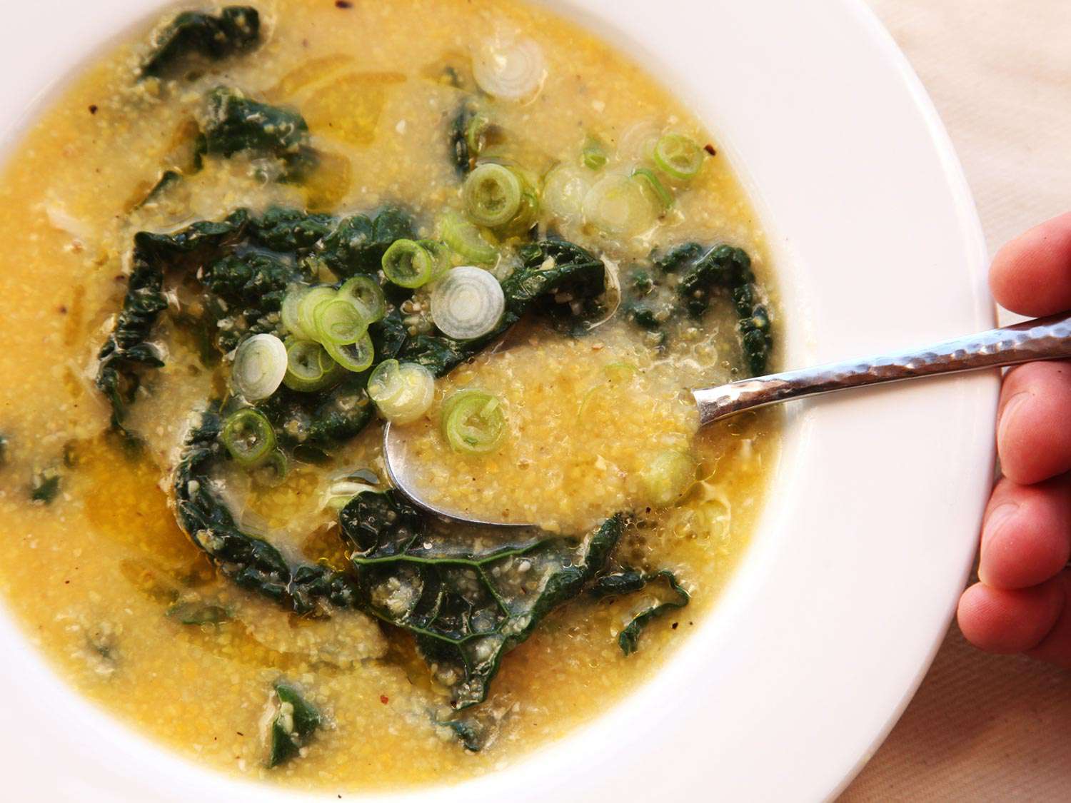 A spoon ladles up some vegan polenta and kale soup from a serving bowl.