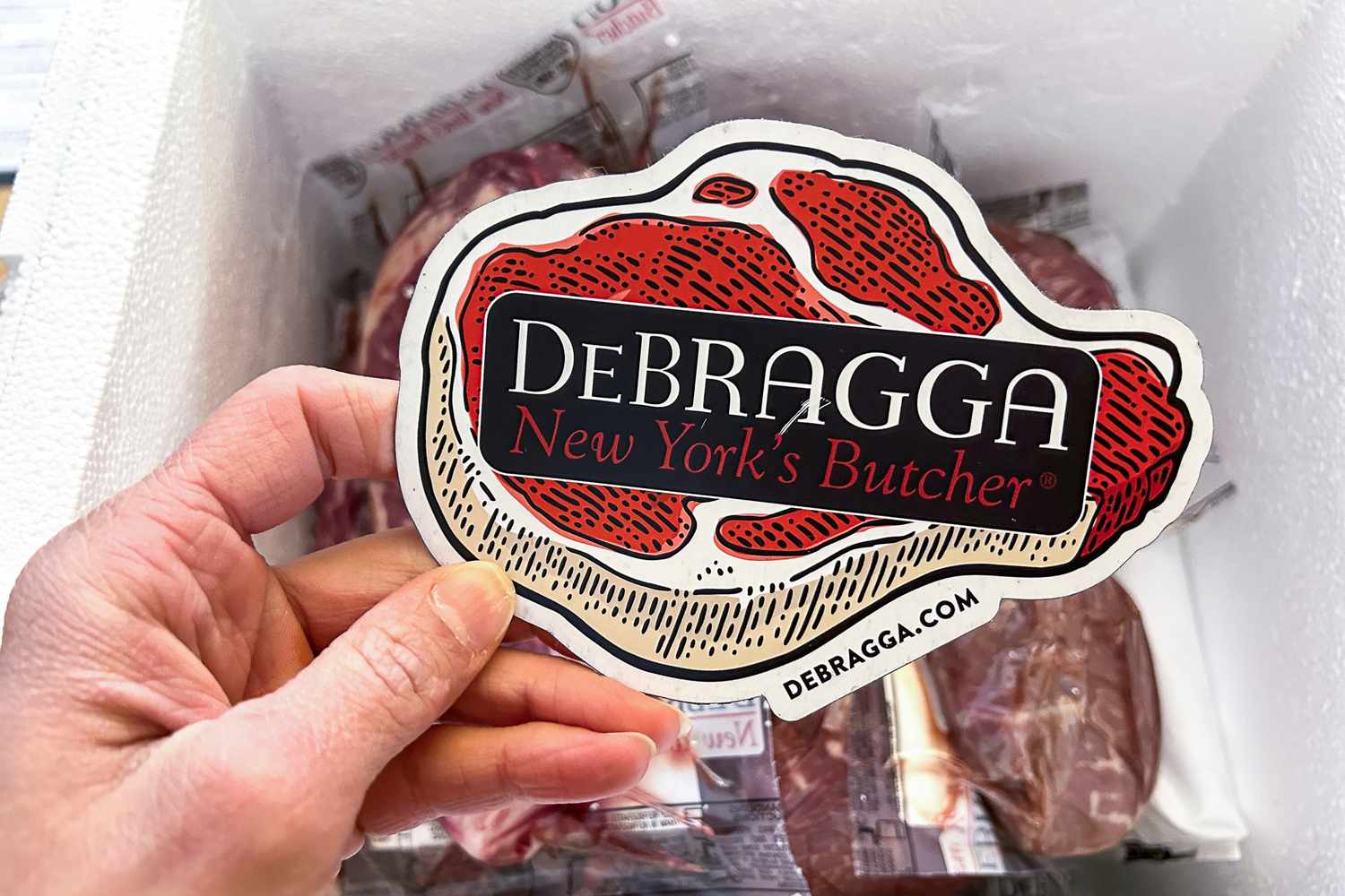 A hand holding a sticker from DeBragga a butcher shop depicting a steak illustration