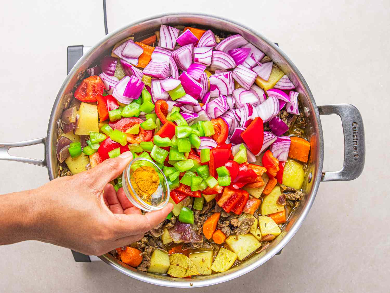 A hand adding spices to a pot containing chopped vegetables and curry ingredients