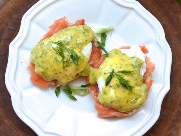 A white plate with two smoked salmon eggs Benedict, garnished with dill hollandaise and herbs. 