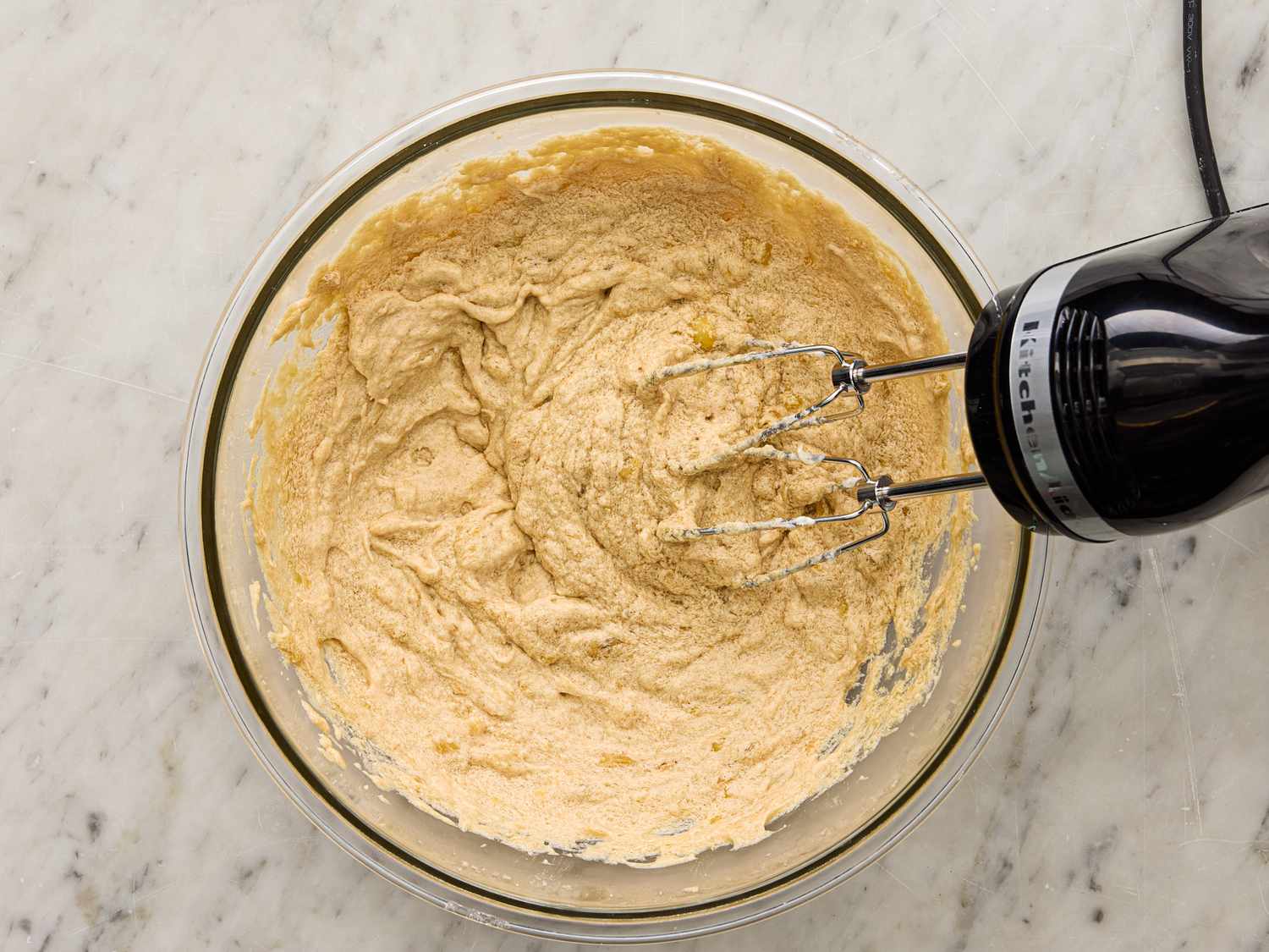 Mixing batter with a hand mixer in a glass bowl on a marble surface