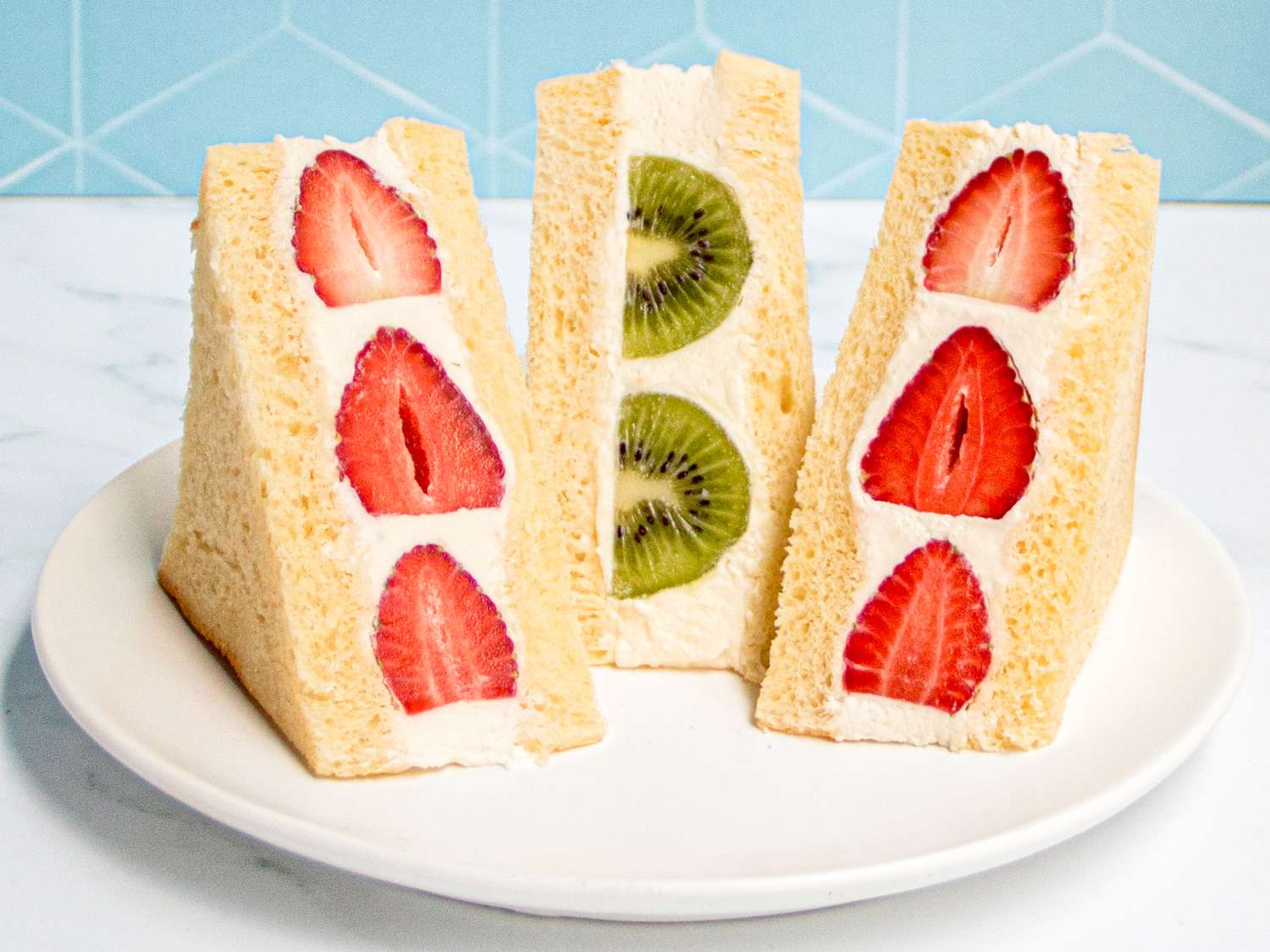 Side view of three fruit sandos, two with strawberries and one with kiwi on a white plate on marble in front of a blue tile wall.