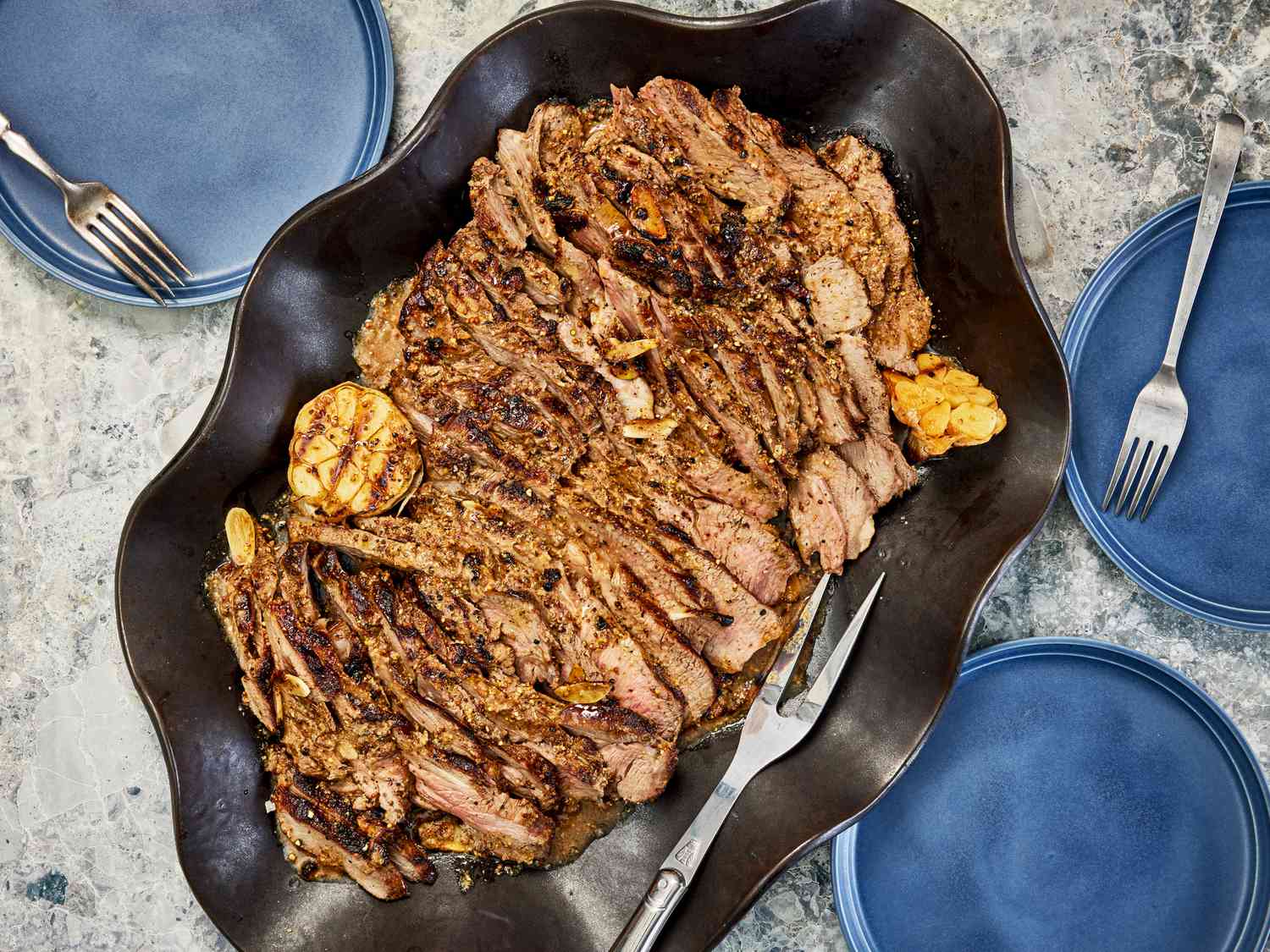 Overhead view of grilled butterflied leg of lamb in a serving platter surrounded by blue plates