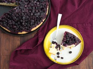 A plated wedge of upside down blueberry muffin cut from a whole cake, with a fork pierced into a piece, 