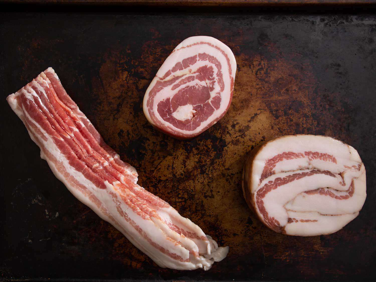 Overhead view of bacon, pancetta, and guanciale arranged on a well-seasoned baking sheet.