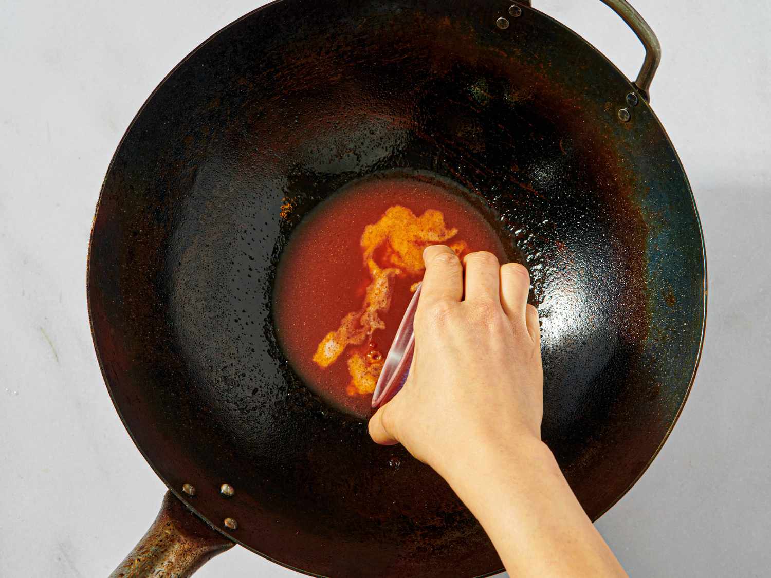 A hand pouring red liquid into a wok preparing a dish on the stove