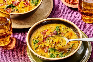 Bowls of pumpkin soup with wild rice and greens spoons placed in one bowl glasses of drink nearby