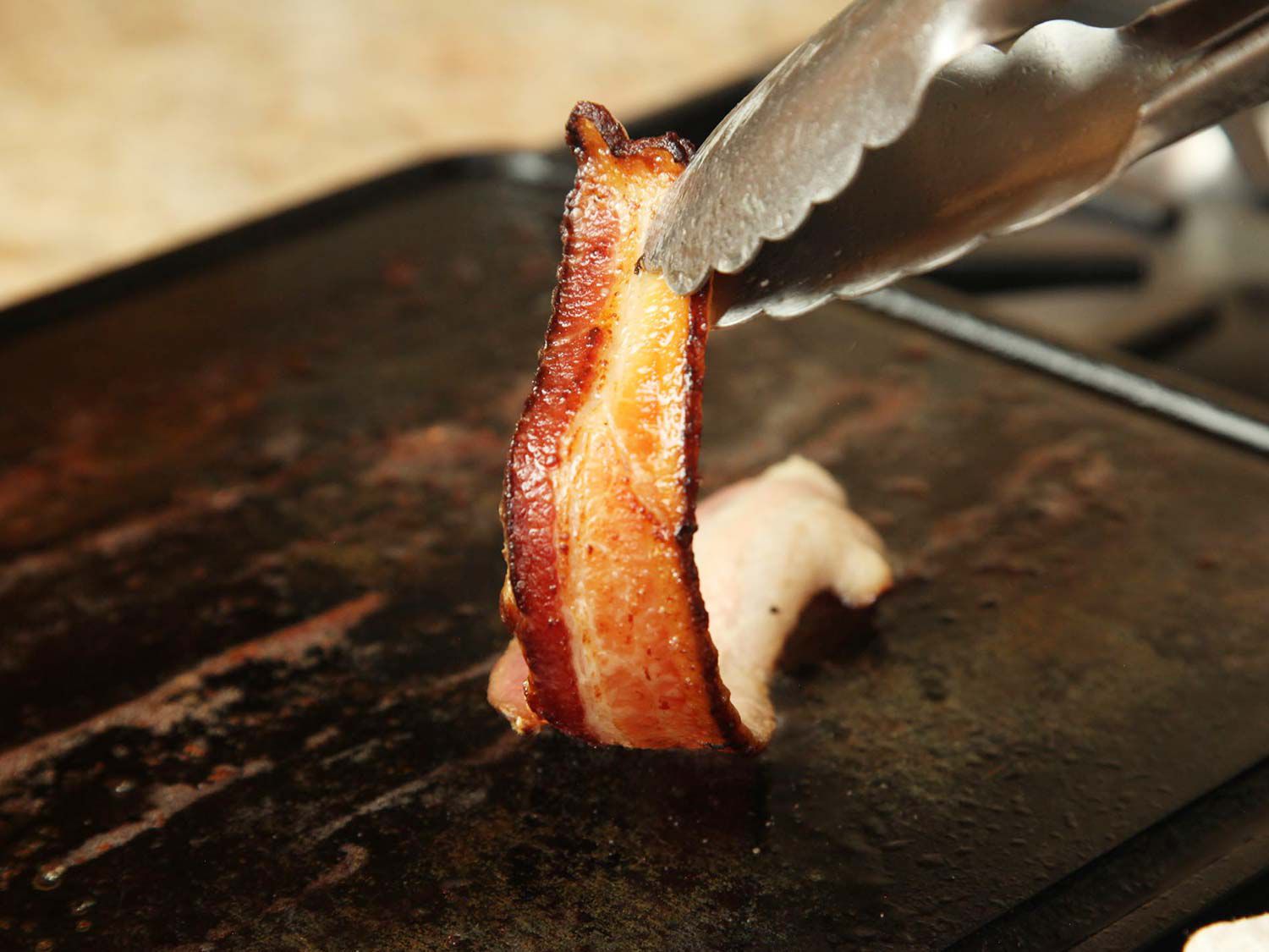Author folding up a slice of bacon with a pair of tongs to reveal the browned, crisped underside.