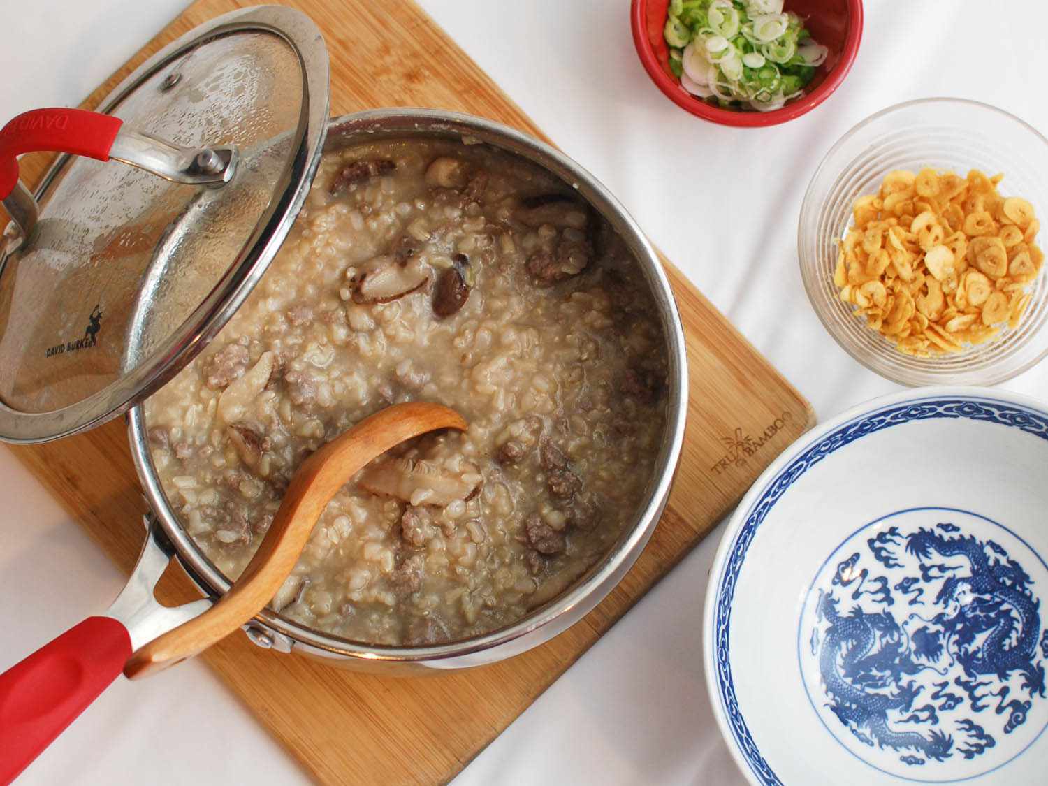 Overhead view of a saucepan with the finished congee, set on a cutting board next to a bowl of the fried garlic chips and a bowl of sliced scallions.