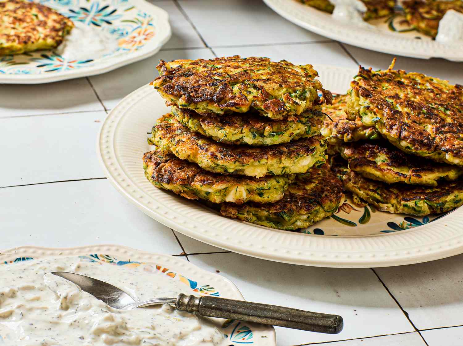 A plate of fritters stacked on a floralpatterned dish served with a side of yogurt sauce and a knife