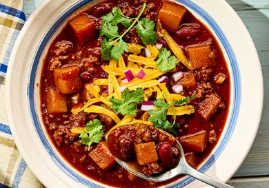 A bowl of turkey chili with squash beans cheese onions and cilantro garnish spoon served alongside