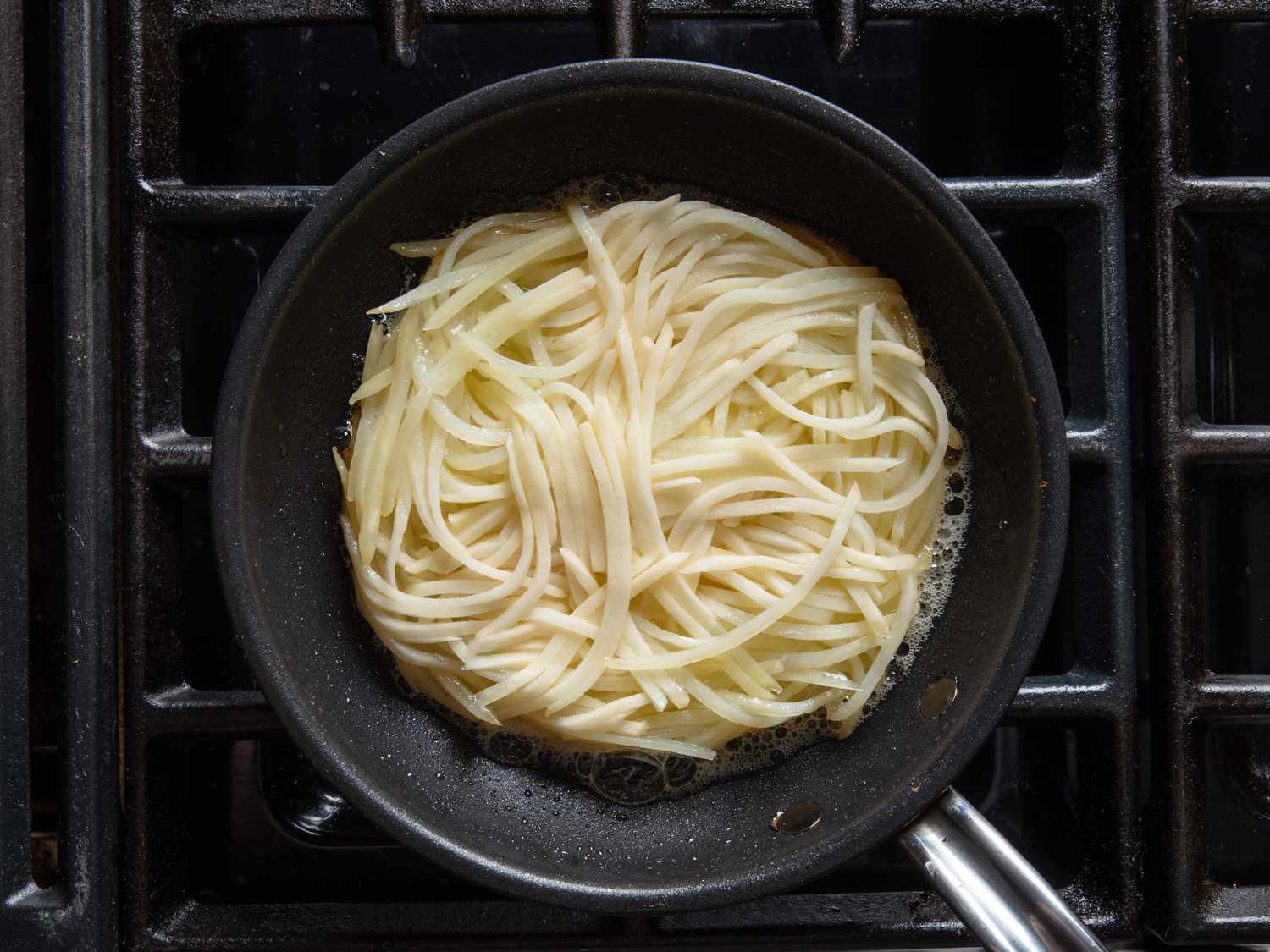 The pommes Darphin is in a skillet, the top side is still raw, but you can see that the potato strands are beginning to soften and hold a more rounded cake shape.