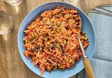 Carrot slaw in a blue bowl surrounded by glasses of water on a wooden surface with napkins and serving spoon 
