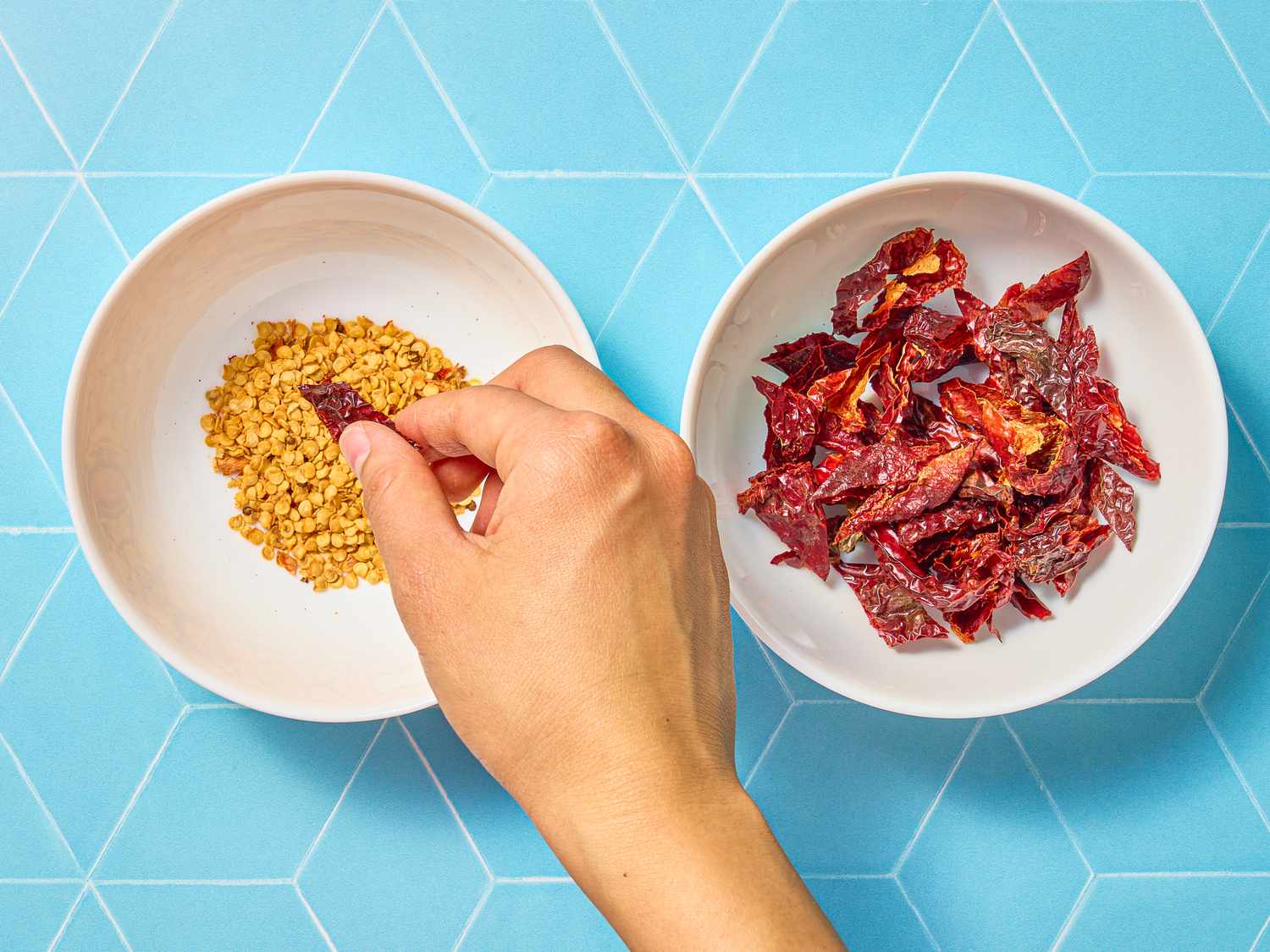 A hand reaching for dried chilies from a bowl next to another bowl containing a different ingredient against a blue patterned background
