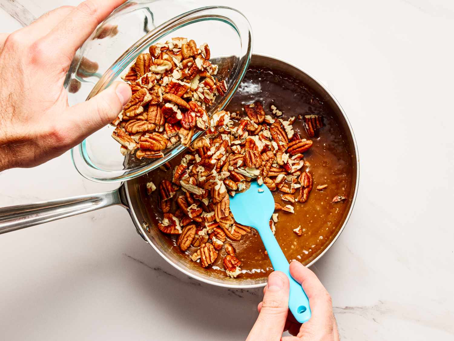 A hand pouring pecans into a saucepan containing a caramel mixture with another hand stirring using a blue spatula