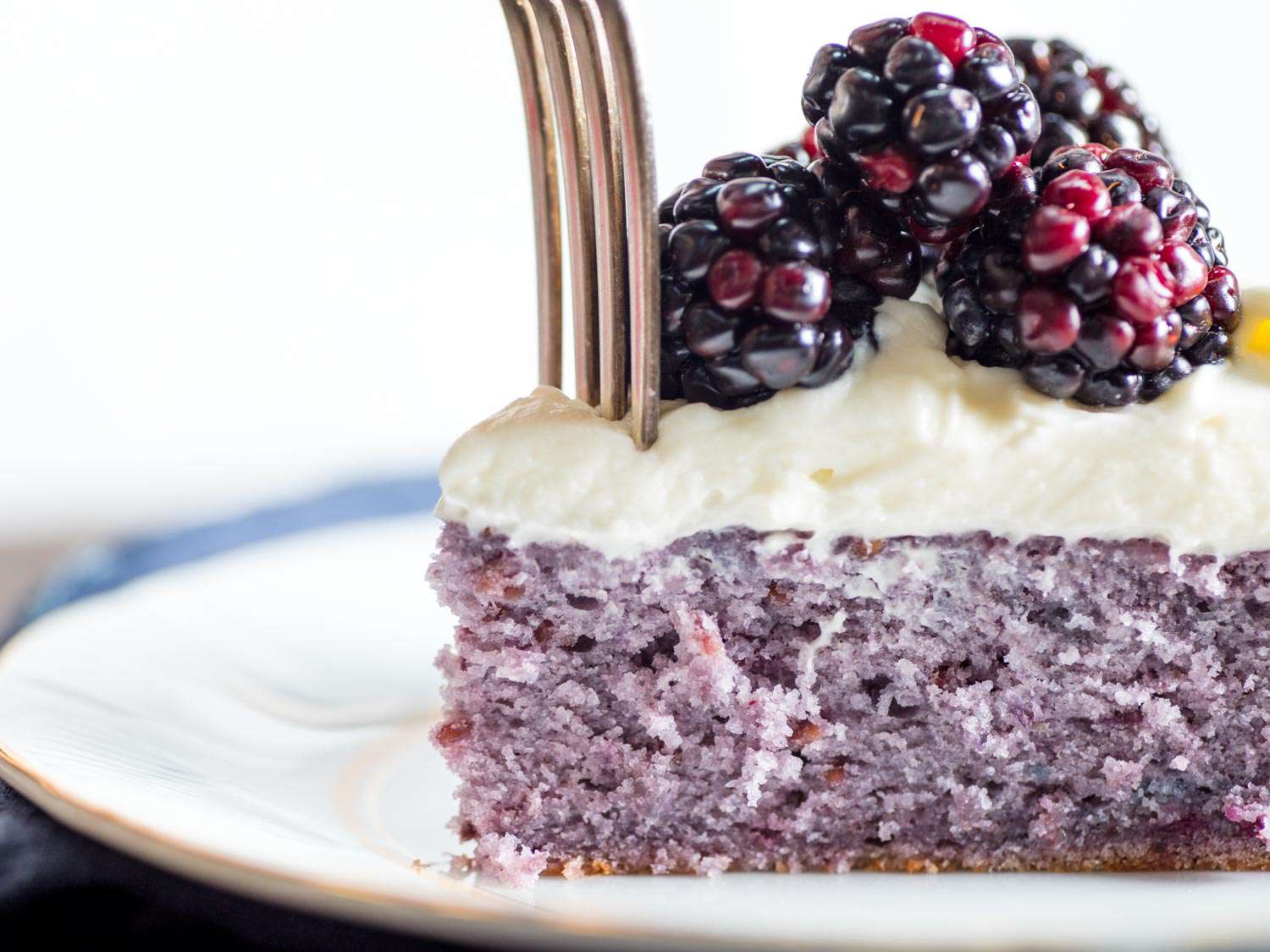 Close-up of a slice of blackberry cake on a plate, topped with cream cheese frosting and fresh blackberries, with a fork sliding in