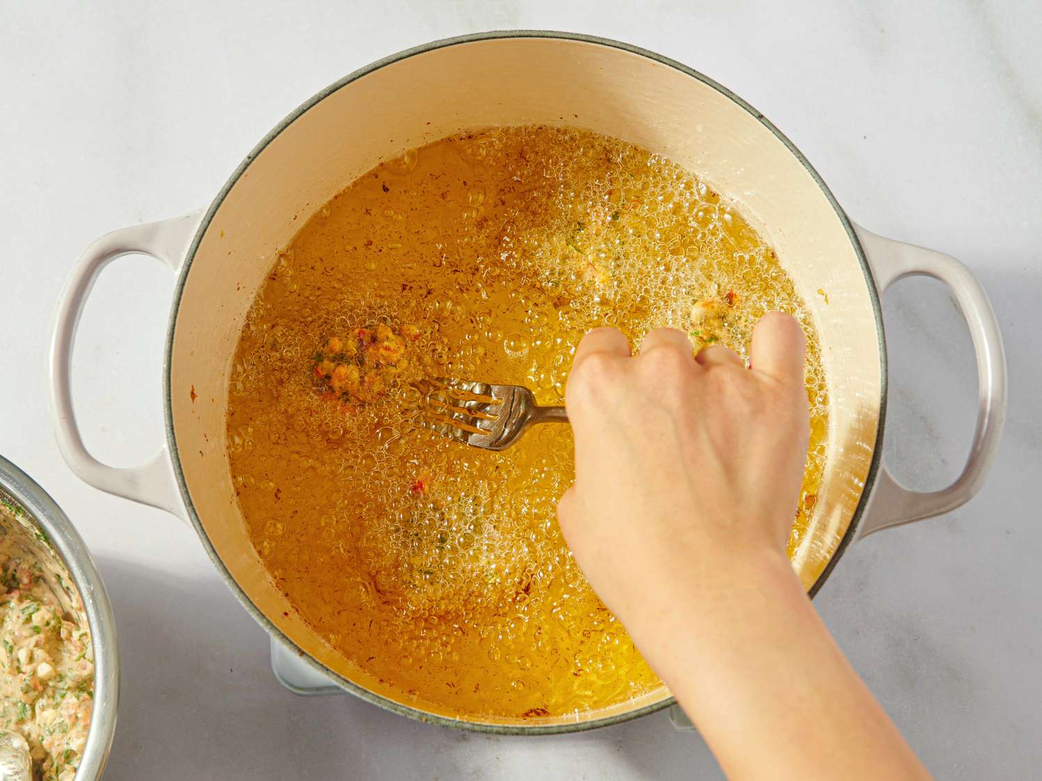 A hand holds a fork over a pot with frying food likely for preparing a dish