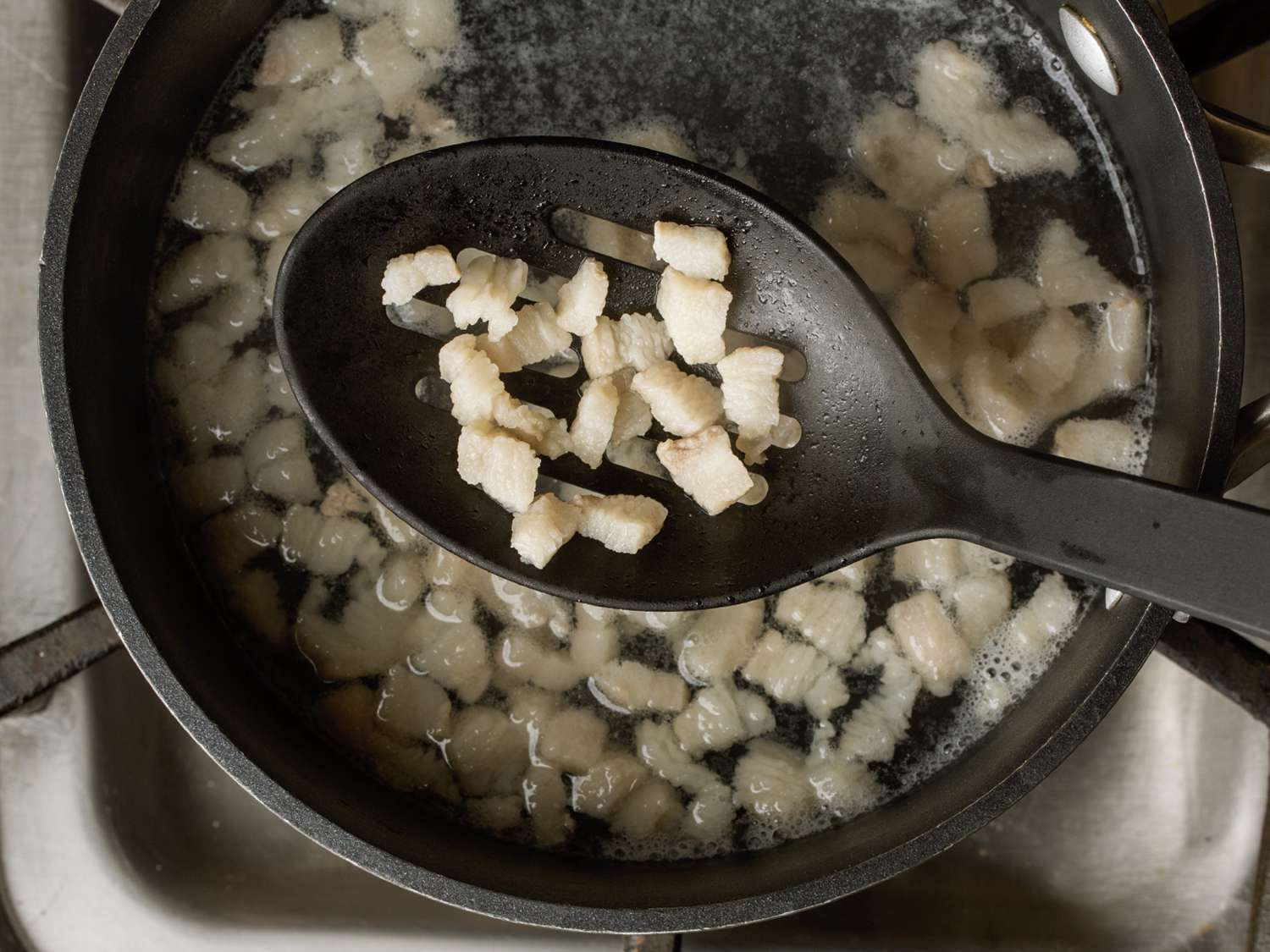 Slotted spoon with pieces of boiled pork fatback held over a pot of boiling water with more pork fatback pieces