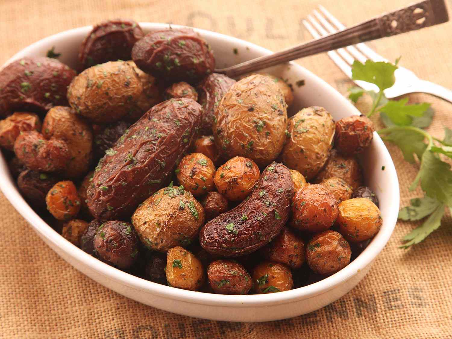 An oval white bowl filled with crispy, herb-flecked roasted potatoes.