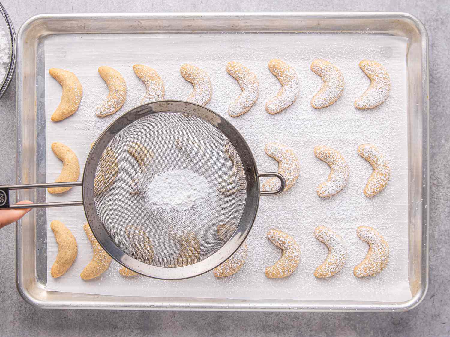 A baking tray of crescentshaped cookies being dusted with powdered sugar using a sieve