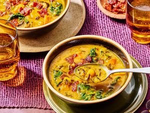 Bowls of pumpkin soup with wild rice and greens spoons placed in one bowl glasses of drink nearby