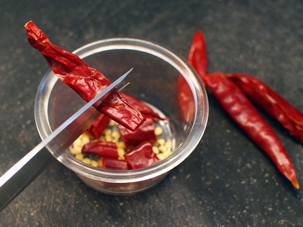 A pair of kitchen shears cutting through a dried chile, overlooking a bowl that catches the chile's seeds and segments. 