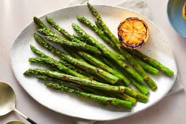 Grilled asparagus served on a large serving plate, drizzled with olive oil, with lemon squeezed over the top, and garnished with charred lemons.