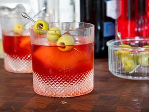 A glass of a cocktail garnished with green olives on a pick placed on a wooden surface with bottles and a dish of olives in the background