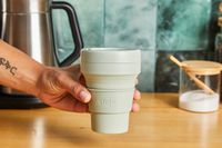 A person picking up a Stojo travel cup off of a ktichen countertop.