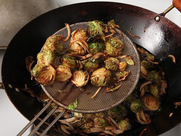 Halved Brussels sprouts and sliced shallots being deep-fried in a wok. A metal spider is holding some of the vegetables above the wok. 