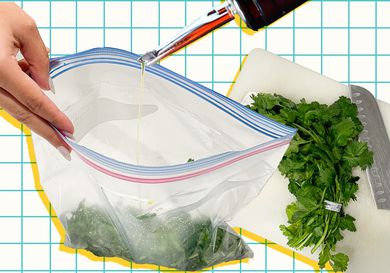 Herb s in a bag being drizzled with olive oil next to a cutting board with a bunch of herbs and a knife. 