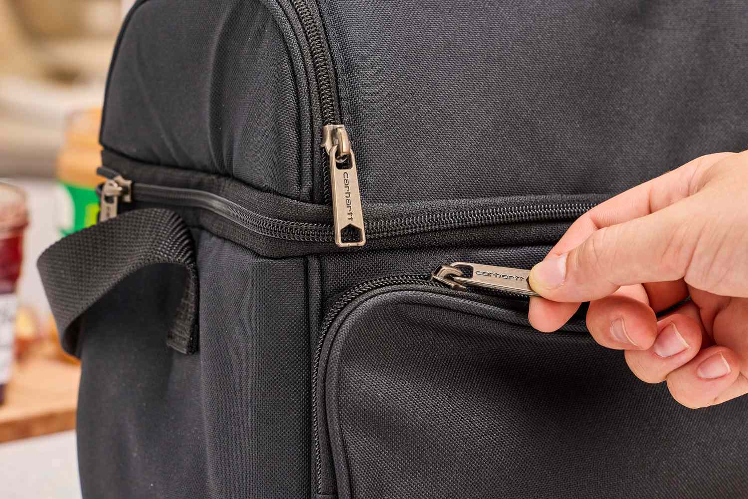 A hand opens a zippered compartment of a black lunch box.