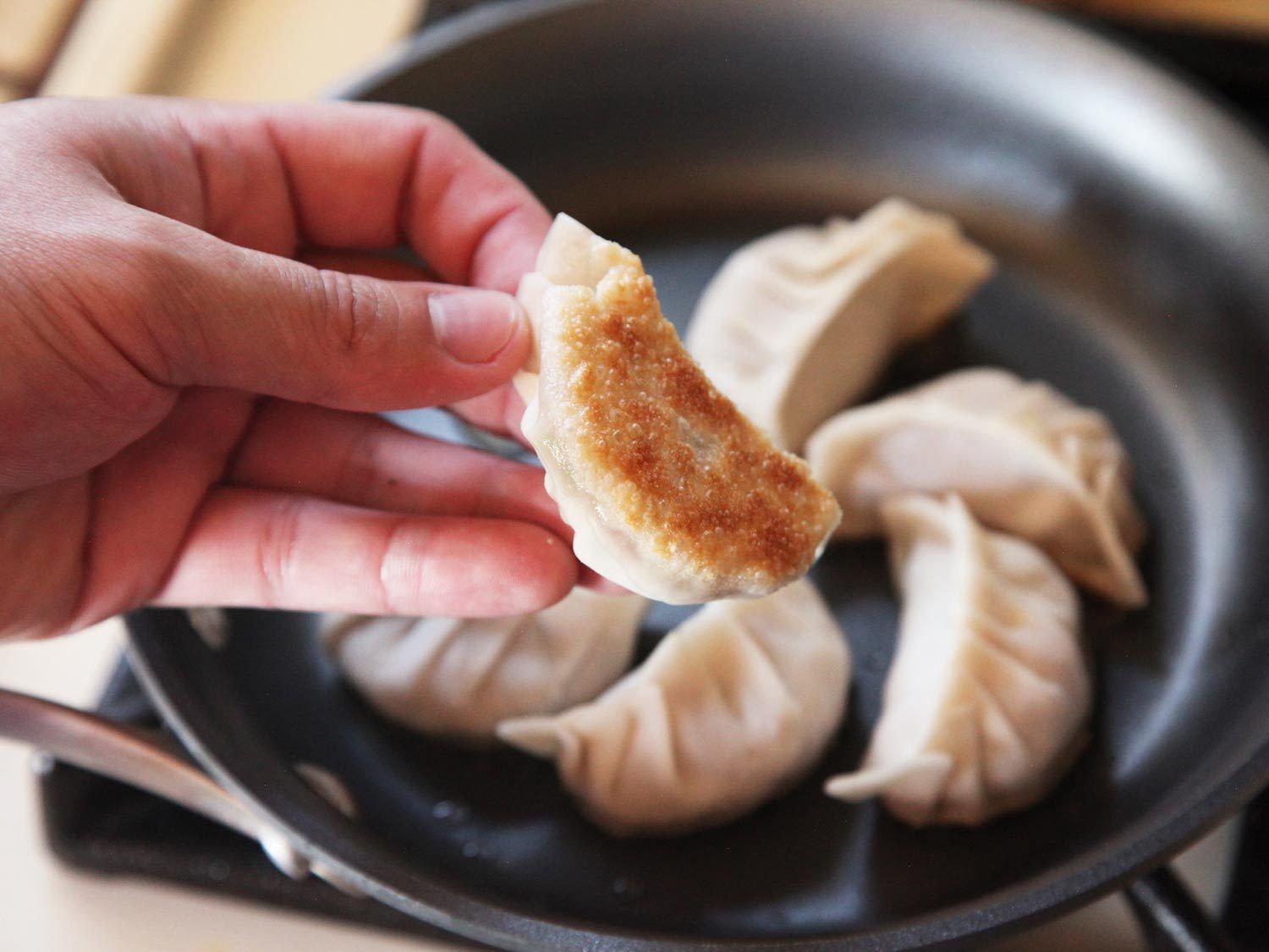 A hand showing the golden-brown bottom of a crisply fried dumpling, more dumplings in a skillet in the background.