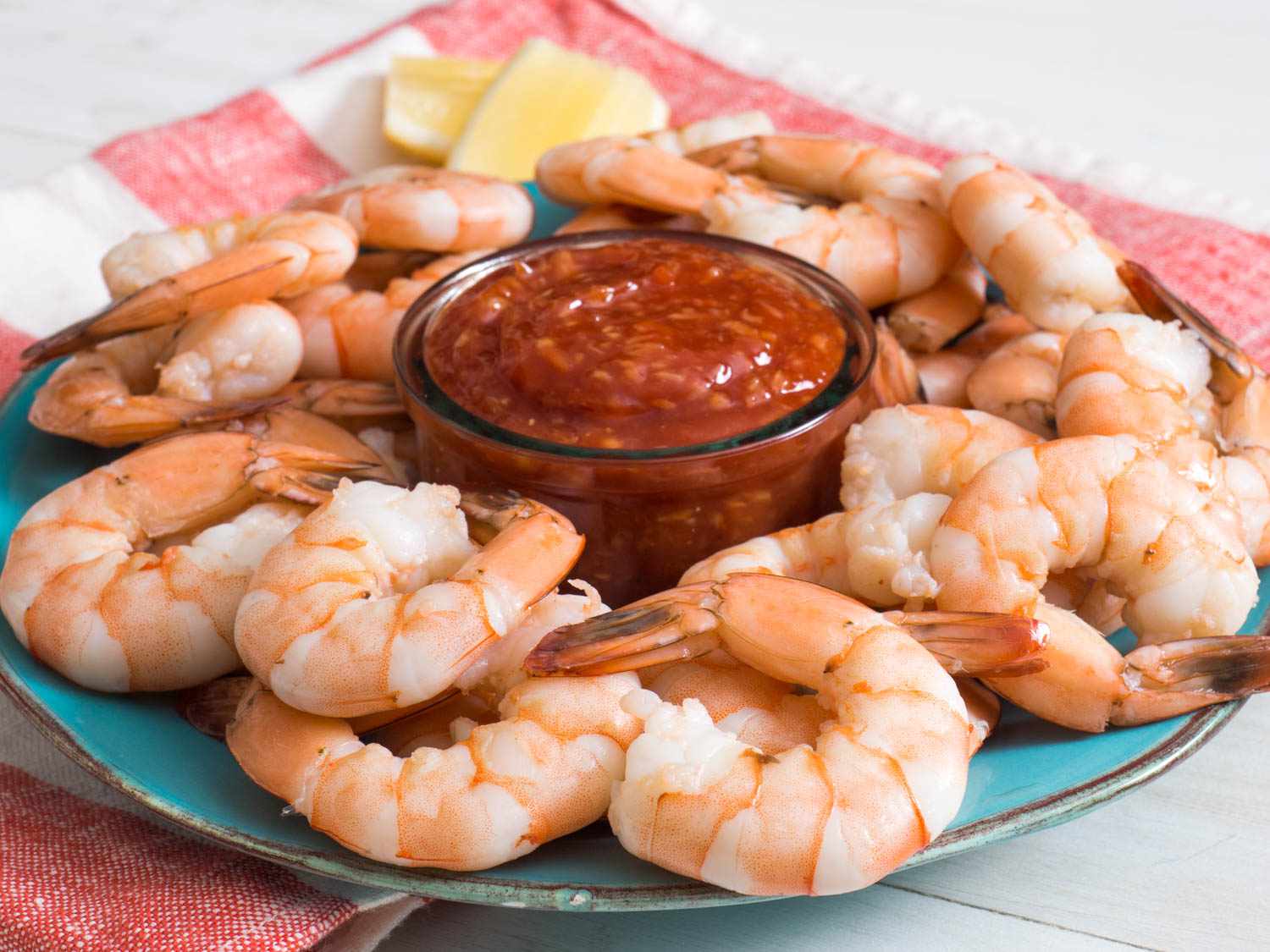 A blue plate covered in plump and tender shrimp with a glass bowl of cocktail sauce at the center. 