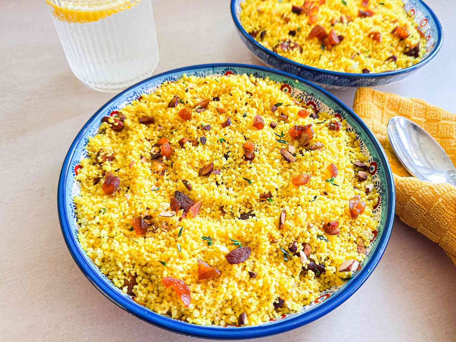 Bowls of couscous garnished with dried fruits and nuts on a table near a drink and a yellow napkin with spoon