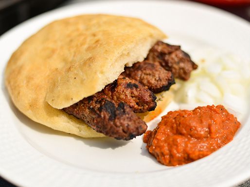 Cevapi sandwiched in Serbian bread with raw onions and roasted red pepper and eggplant sauce on the side .