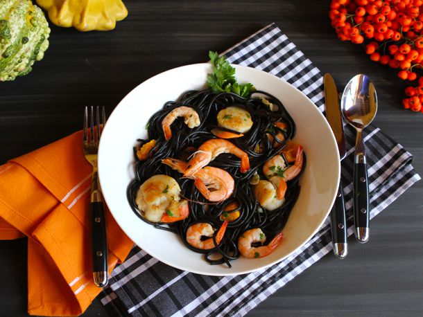 A bowl of squid ink pasta with shrimp and scallops, flanked by silverware and an orange napkin.